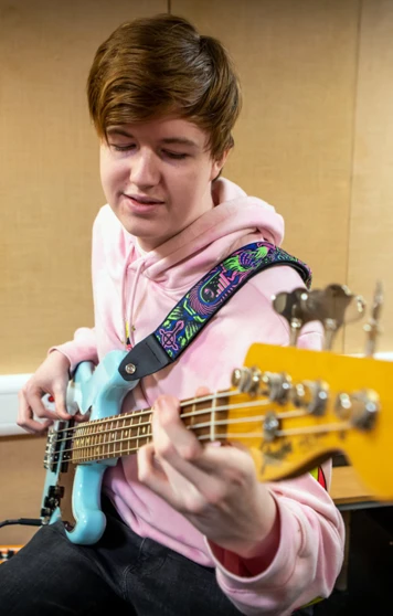 A young student in a pink hoodie playing a blue bass guitar in a music practice room, focusing on their instrument. A young student in a pink hoodie playing a blue bass guitar in a music practice room, focusing on their instrument.